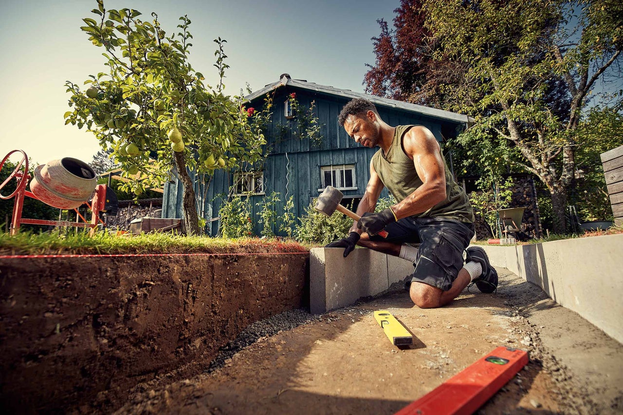 Ein Mann bearbeitet einen Pflasterstein mit einem Hammer im Garten.
