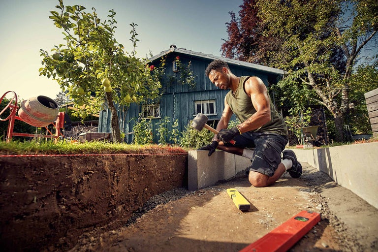 Ein Mann bearbeitet einen Pflasterstein mit einem Hammer im Garten.