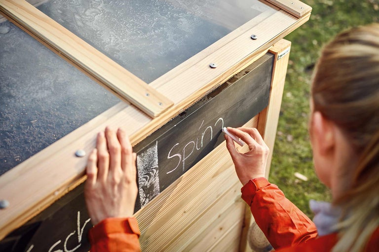 Nahaufnahme eines Frühbeets aus Holz mit einer Person, die mit Kreide "Spinat" auf eine Tafel schreibt.