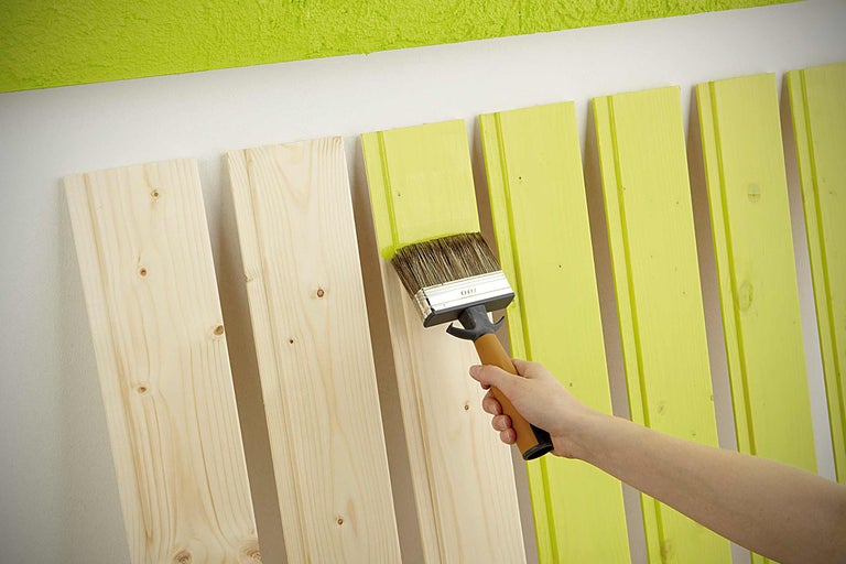 Eine Person streicht Holzlatten mit einem Pinsel in einem hellen Farbton.