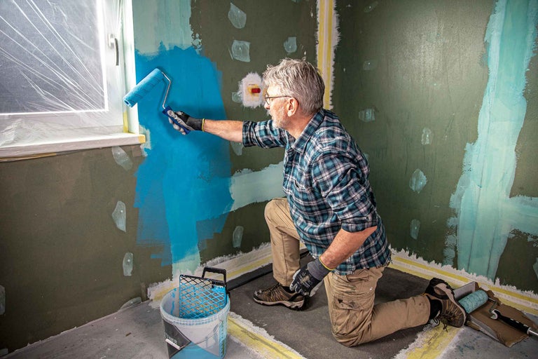 Ein Mann streicht eine Wand mit einer Farbrolle in einem Raum. Fenster und Boden sind mit Folie abgedeckt.