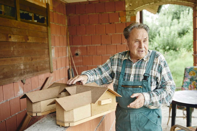 Ein Mann im Blaumann zeigt auf mehrere kleine Modellhäuser aus Holz vor einer Ziegelwand.