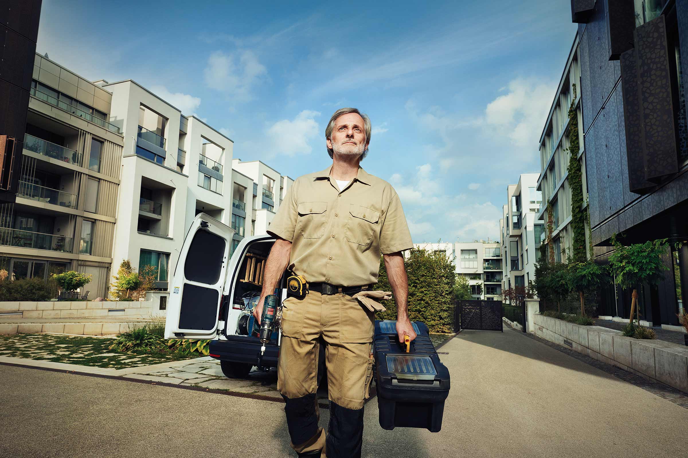 Handwerker mit Werkzeugkiste und Bohrmaschine vor einem Lieferwagen und Wohnhäusern