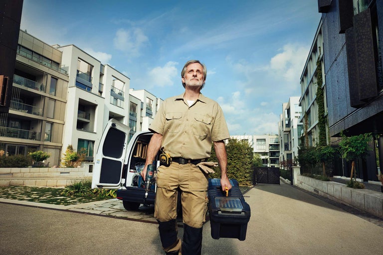 Handwerker mit Werkzeugkiste und Bohrmaschine vor einem Lieferwagen und Wohnhäusern