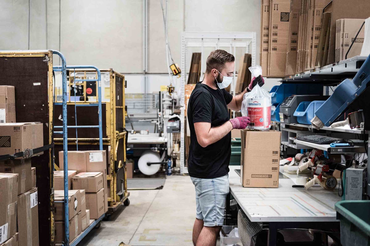 Ein Mitarbeiter im Lager verpackt Remmers Holzlasur in einem Karton für den Versand.