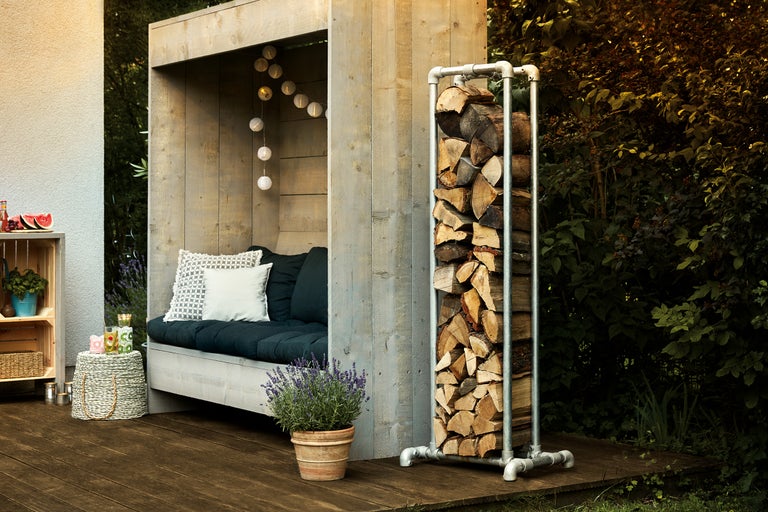 Holzlager aus Metall mit Holzscheiten neben einer Holzhütte mit Sitzgelegenheit auf einer Terrasse