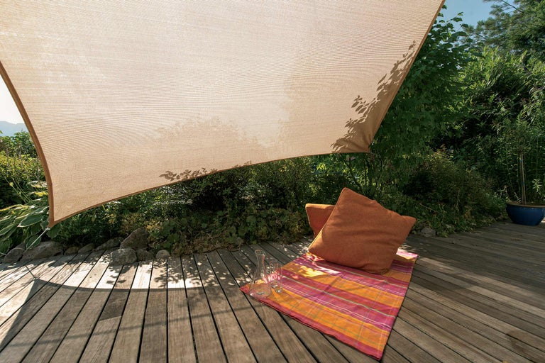 Sonnensegel auf einer Holzterrasse mit Kissen und Decke im Garten.