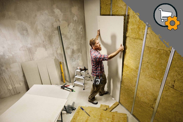 Ein Mann montiert eine Gipskartonplatte auf einer Wand mit Dämmmaterial und Metallprofilen. Auf dem Boden liegen weitere Platten und Werkzeuge.