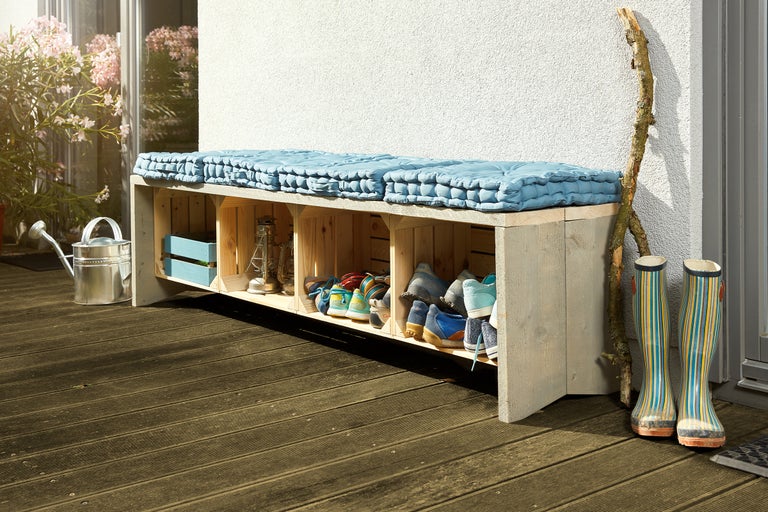Garderobenbank aus Holzkisten mit Schuhablage und Sitzkissen auf einer Terrasse mit Gießkanne und Gummistiefeln