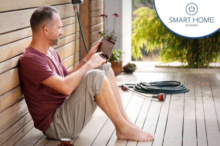 Mann sitzt mit Tablet auf einer Holzterrasse; Gartenschlauch im Hintergrund; Hornbach Smart Home Logo