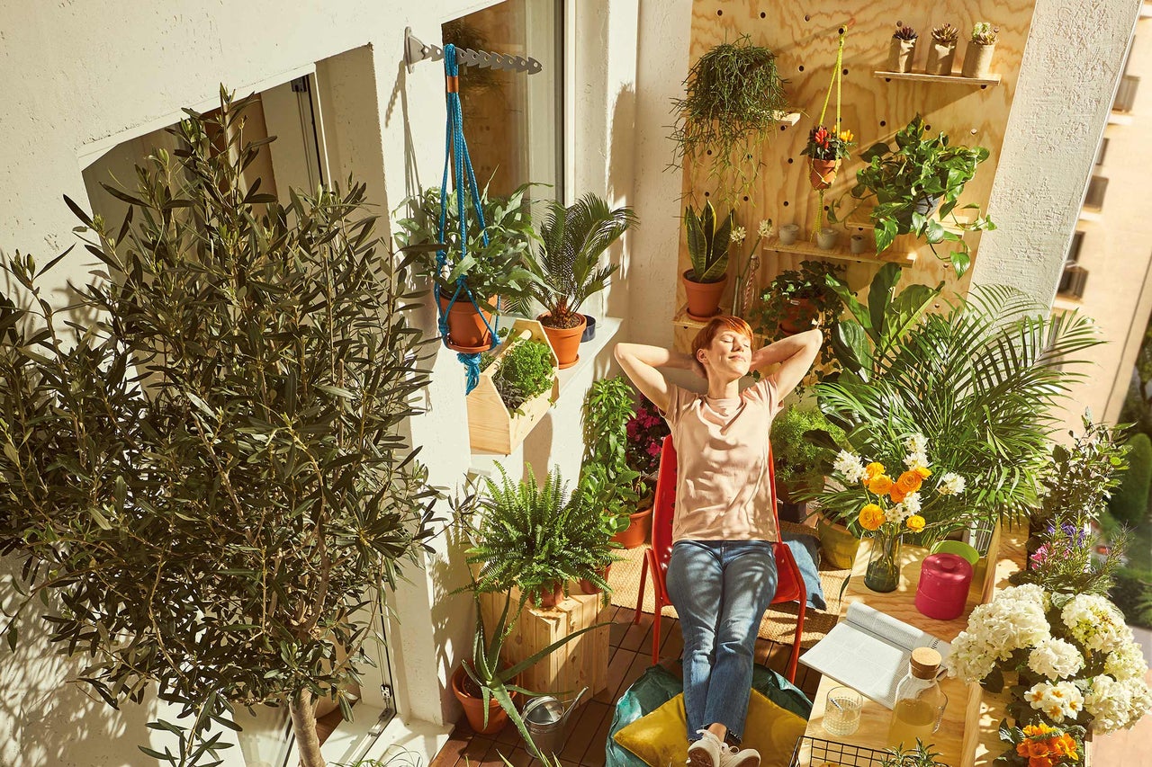 Frau entspannt auf einem sonnigen Balkon, umgeben von Pflanzen in Töpfen und hängenden Pflanzgefäßen.