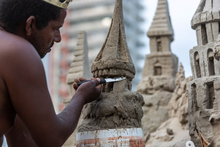 Ein Mann mit Krone formt eine Sandburg mit einem Messer.