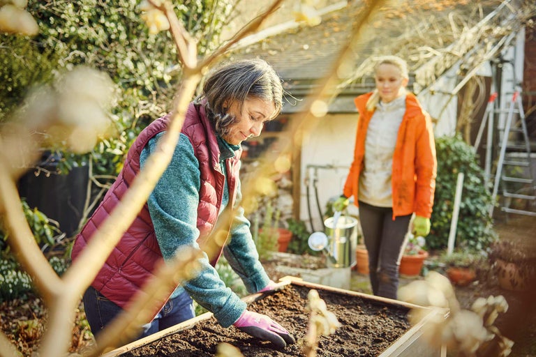 Gartenarbeitsszene mit zwei Frauen, einer mit Gartenhandschuhen in einem Hochbeet, die andere mit einer Gießkanne im Hintergrund.
