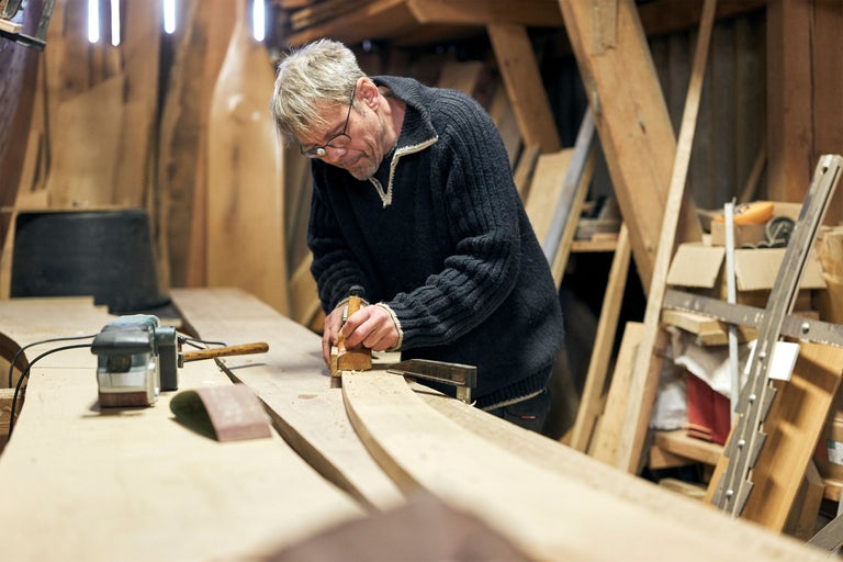 Ein Handwerker bearbeitet ein Holzstück mit einem Hobel in einer Werkstatt. Werkzeuge und Holz sind im Hintergrund zu sehen.