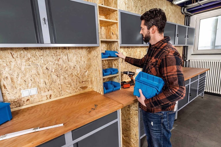 Mann ordnet Werkstatt mit blauen Lagerboxen in einem OSB-Regal über einer Holzwerkbank mit grauen Werkstattschränken an.