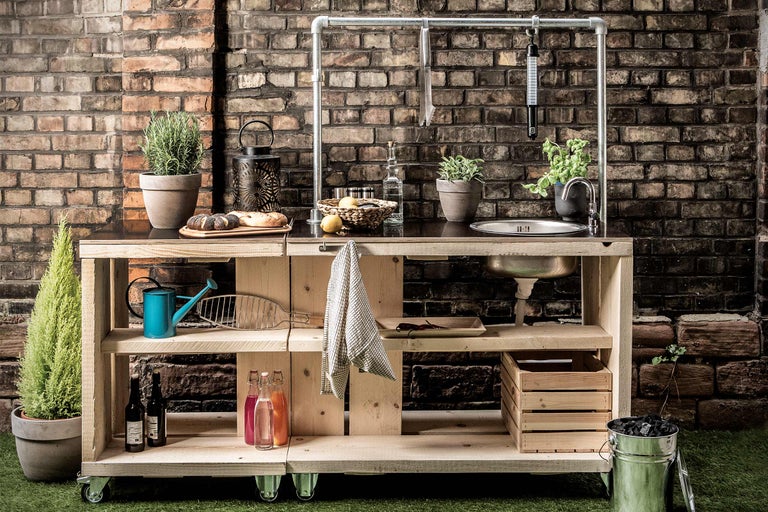 Outdoor-Küche mit Holzkonstruktion, Spüle, Ablageflächen und Rädern vor einer Backsteinmauer