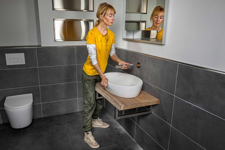Badezimmer mit Keramikwaschtisch, Holztischplatte und Armaturinstallation durch eine Frau