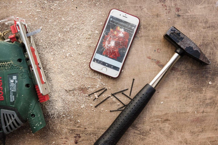 Szene mit Stichsäge, Hammer, Nägeln und einem Smartphone mit gesprungenem Display auf einer Werkbank