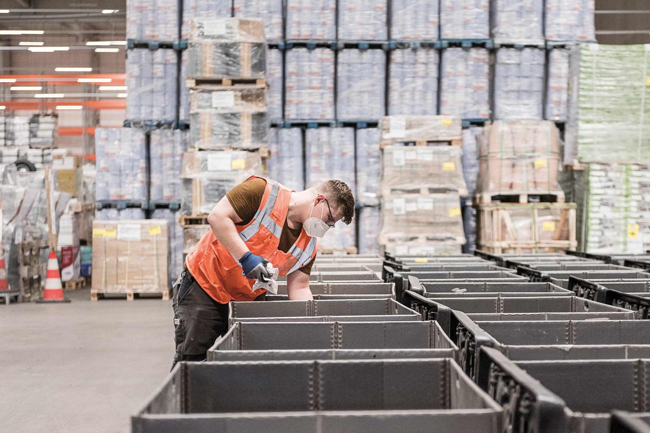 Ein Arbeiter mit Schutzweste und Mundschutz in einem Lagerhaus mit grauen Lagerbehältern und Paletten mit Waren.