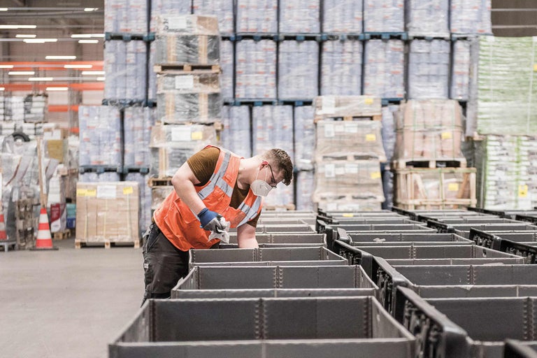 Ein Arbeiter mit Schutzweste und Mundschutz in einem Lagerhaus mit grauen Lagerbehältern und Paletten mit Waren.