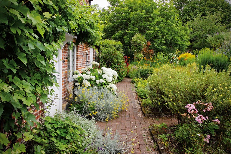 Gepflegter Garten mit Blumen, Büschen und Ziegelweg neben einem Backsteinhaus