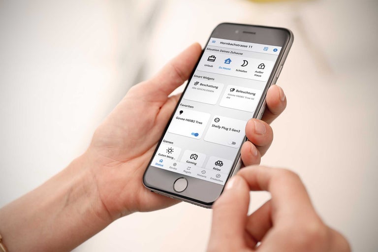 Eine Person bedient eine Smart Home App auf einem Smartphone zur Steuerung von Beleuchtung, Beschattung und verschiedenen Szenarien.