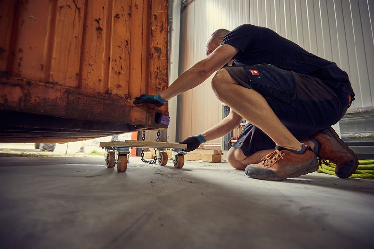 Mann bewegt einen Container mit einem Möbelroller in einer Halle.