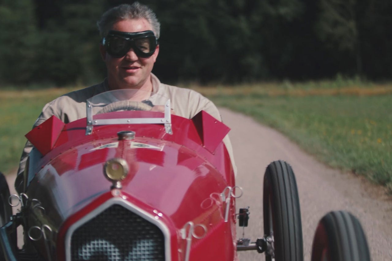 Mann mit Schutzbrille fährt ein rotes Oldtimer-Rennauto auf einer Landstraße