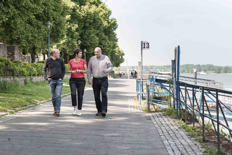 Drei Personen spazieren auf einem Gehweg entlang des Flusses.