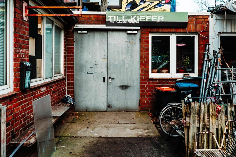 Außenansicht eines Werkstattgebäudes mit Ziegelfassade, einer grauen Metalltür, Fenstern und einem Schild mit der Aufschrift Till Kiefer Metallgestaltung