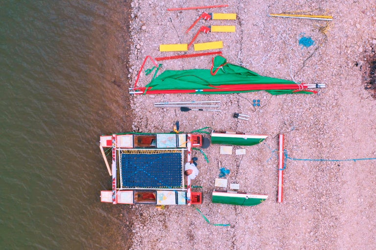 Luftaufnahme von einem zusammengebauten Floß neben einem Flussufer mit dazugehöriger Ausrüstung