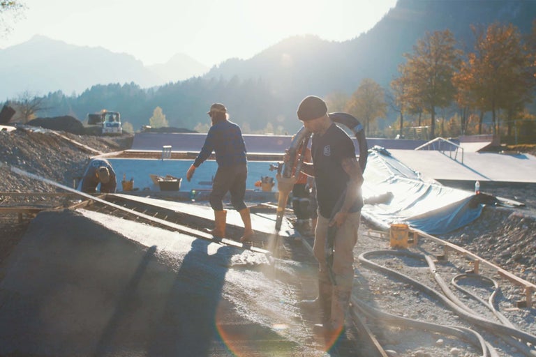 Bauarbeiter beim Betonieren eines Skateparks mit Bergpanorama im Hintergrund