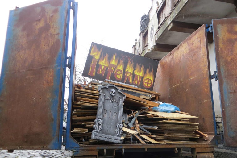 Müllcontainer gefüllt mit Holzplatten und Requisiten, darunter ein Grabstein und ein Schild mit der Aufschrift Inferno.
