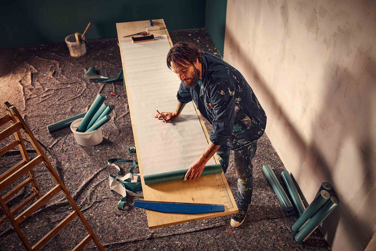 Ein Mann bereitet eine Tapete auf einem Tisch vor, um sie an die Wand zu bringen. Werkzeuge wie eine Leiter, ein Eimer und ein Tapetenroller sind im Raum verteilt.