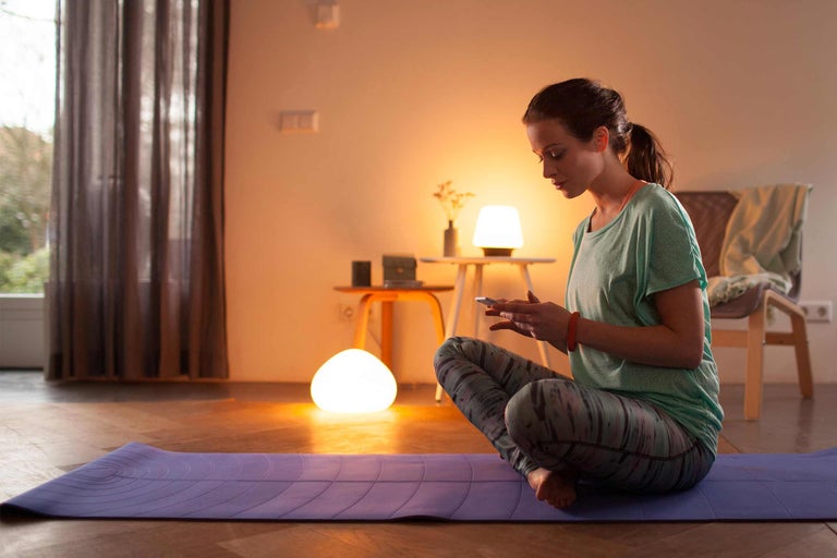Frau sitzt auf einer Yogamatte und benutzt ein Mobiltelefon, Beleuchtung im Raum durch Tischleuchten.
