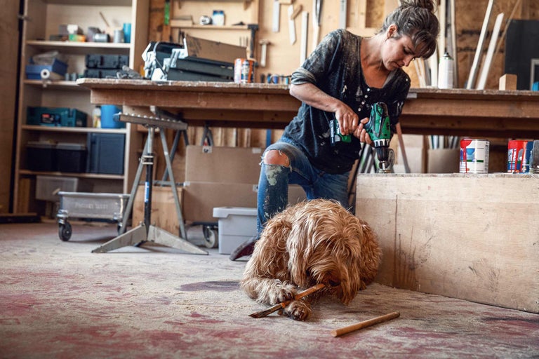 Frau arbeitet mit Bohrmaschine in Werkstatt, Hund liegt im Vordergrund