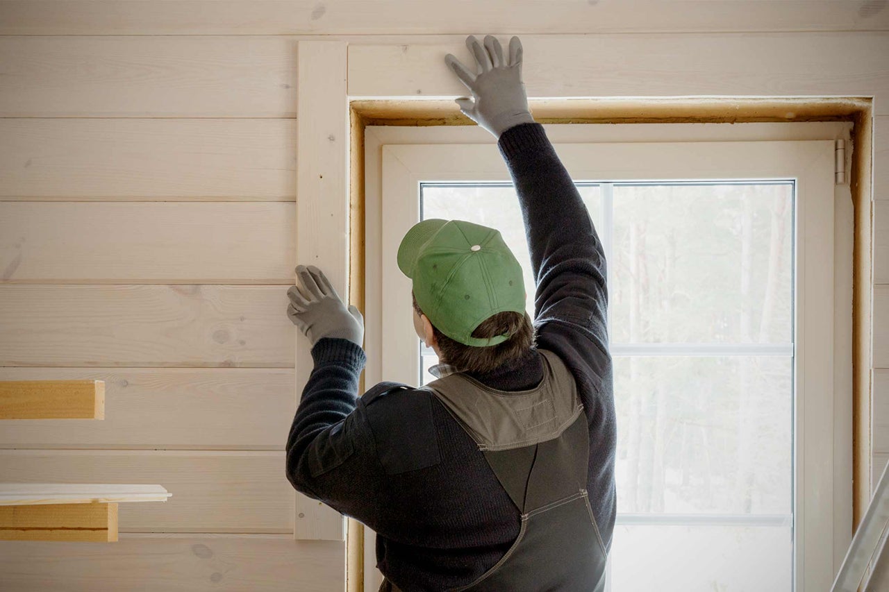 Handwerker montiert Holzrahmen um ein Fenster in einem Raum mit Holzvertäfelung.