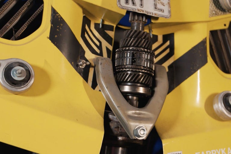 Detailaufnahme eines gelben Fahrzeugbauteils mit Zahnrädern und silberfarbener Halterung.