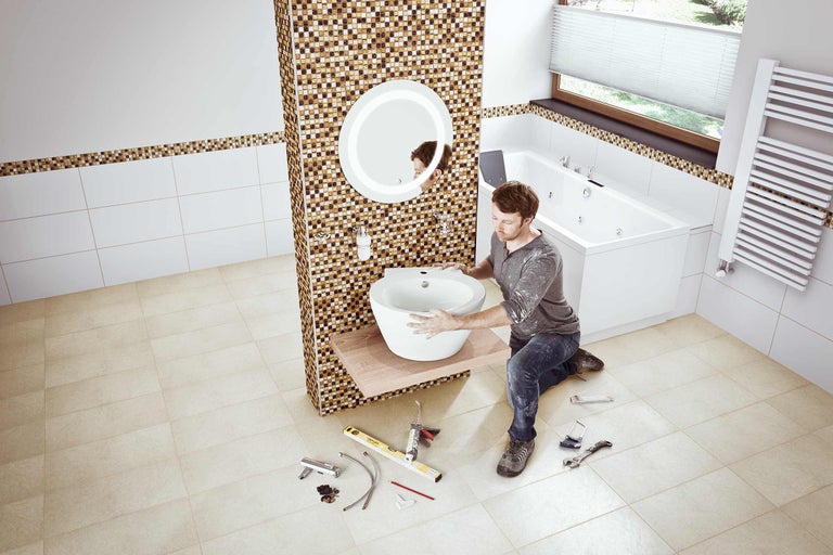 Ein Mann installiert ein Aufsatzwaschbecken in einem Badezimmer mit Mosaikfliesen, Badewanne und Heizkörper.
