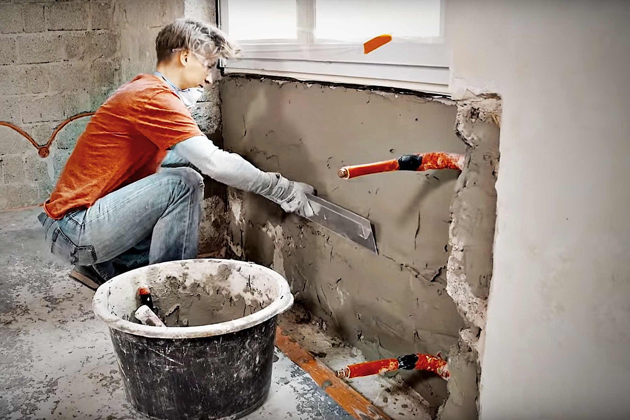 Handwerker trägt Putz auf eine Wand auf, Rohre sind sichtbar.