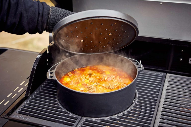 Ein Dutch Oven mit Deckel steht auf einem Grill, gefüllt mit einem Gericht, aus dem Dampf aufsteigt.