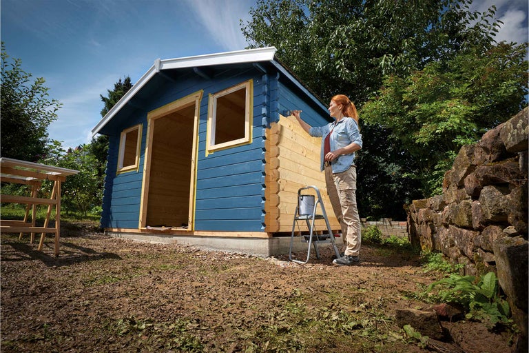 Frau streicht ein Gartenhaus aus Holz mit Farbe
