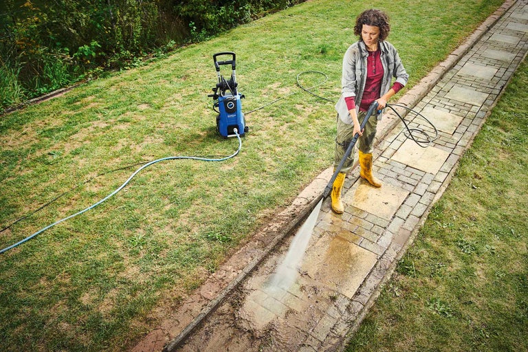 Eine Frau reinigt mit einem Hochdruckreiniger einen Gehweg in einem Garten.