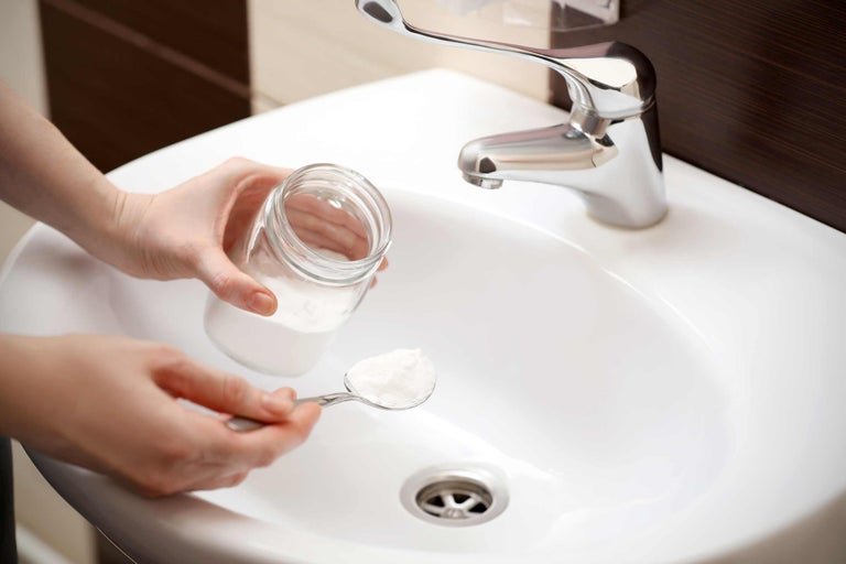 Person hält Glasbehälter und Löffel mit Reinigungspulver über ein Waschbecken mit Wasserhahn