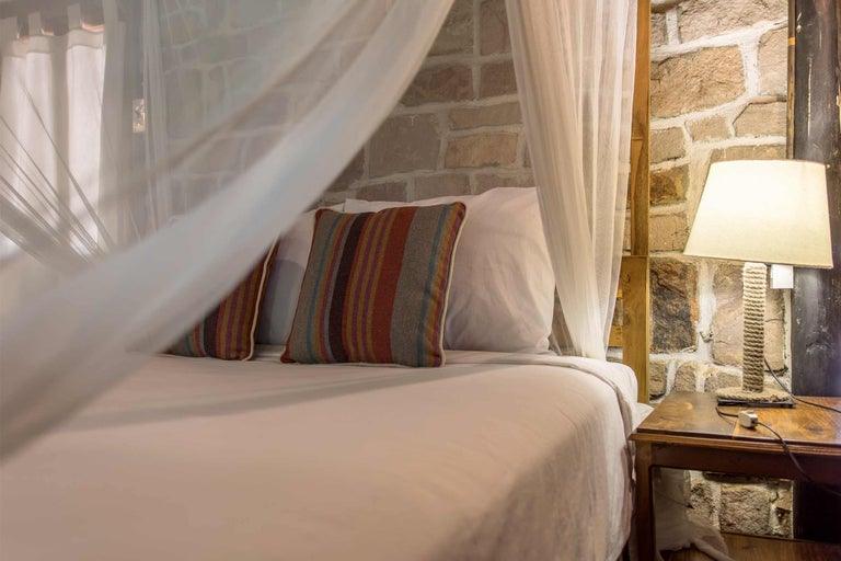 Bett mit Moskitonetz, Kopfkissen und Nachttischlampe vor Steinwand