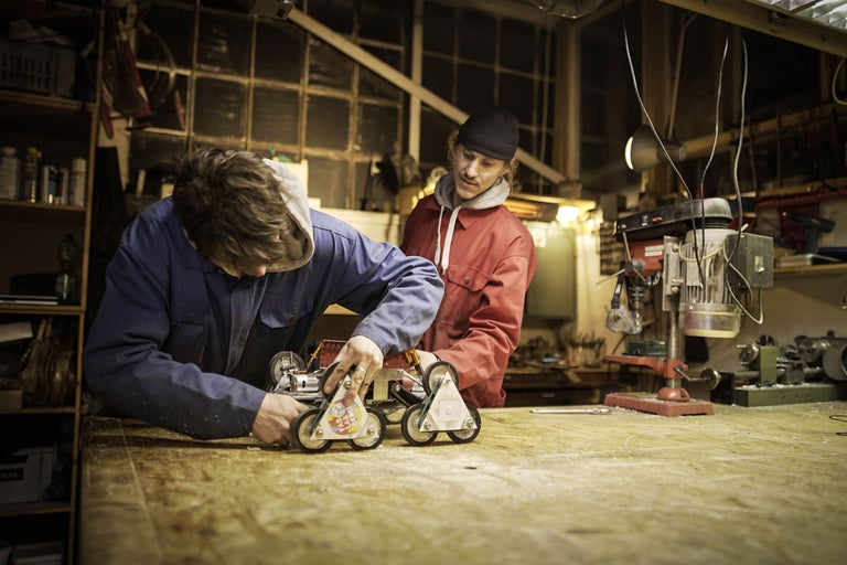 Zwei Männer arbeiten in einer Werkstatt an einem kleinen Gefährt mit Rädern.