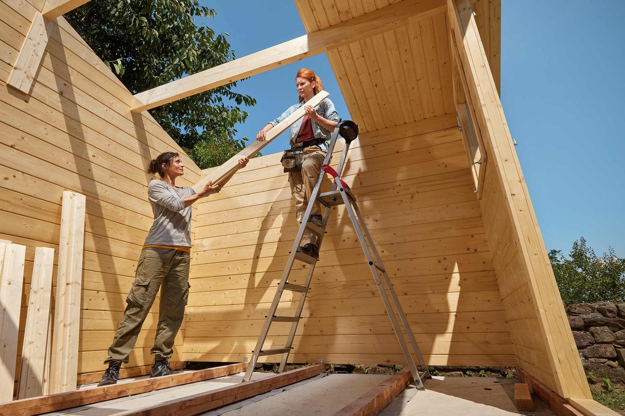 Zwei Personen bauen ein Holzhaus mit einer Leiter.