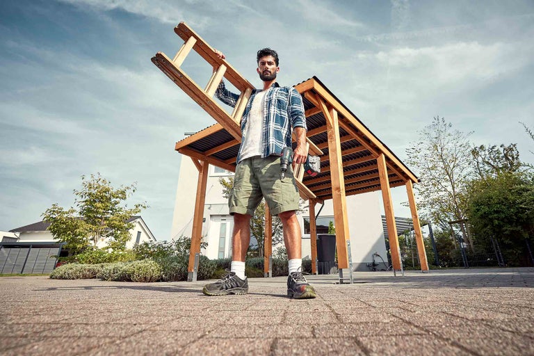 Mann mit Leiter und Akkuschrauber vor einem Carport