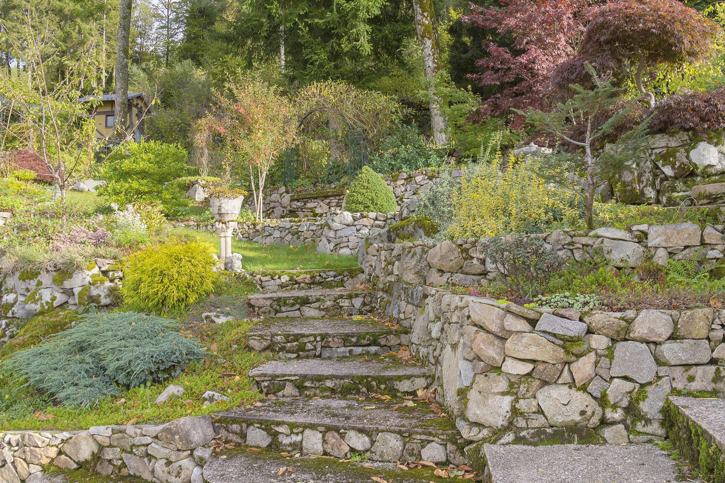 Eine Steintreppe führt durch einen terrassenförmig angelegten Garten mit Steinmauern und verschiedenen Pflanzen