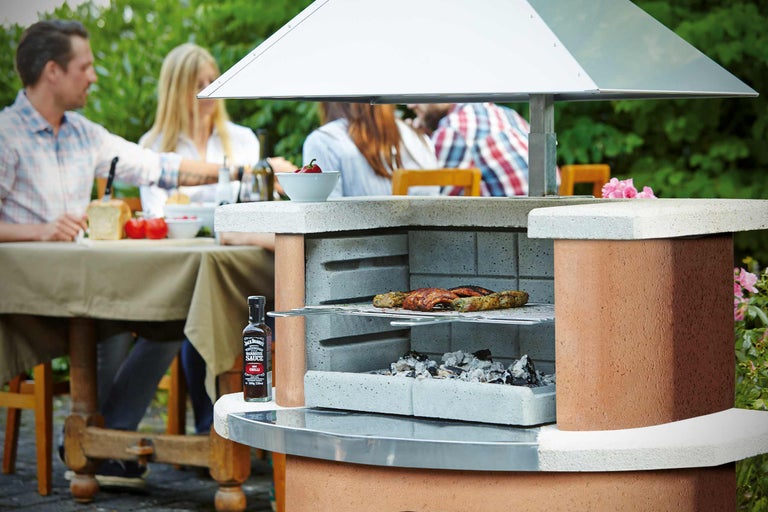 Gemauerter Grill mit Grillgut und Holzkohle im Garten, mit einer Gruppe von Personen, die an einem Tisch sitzen.